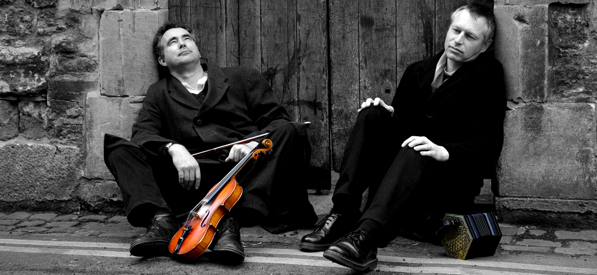Black and white photo Mat Green and Andy Turner sitting by the roadside, only their fiddle and concertina are in colour